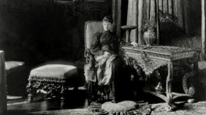 A black and white image of a woman seated next to an ornate table and a cushioned stool. There is a curtained bed in the background and a cushion laying on the patterned carpet on the floor.
